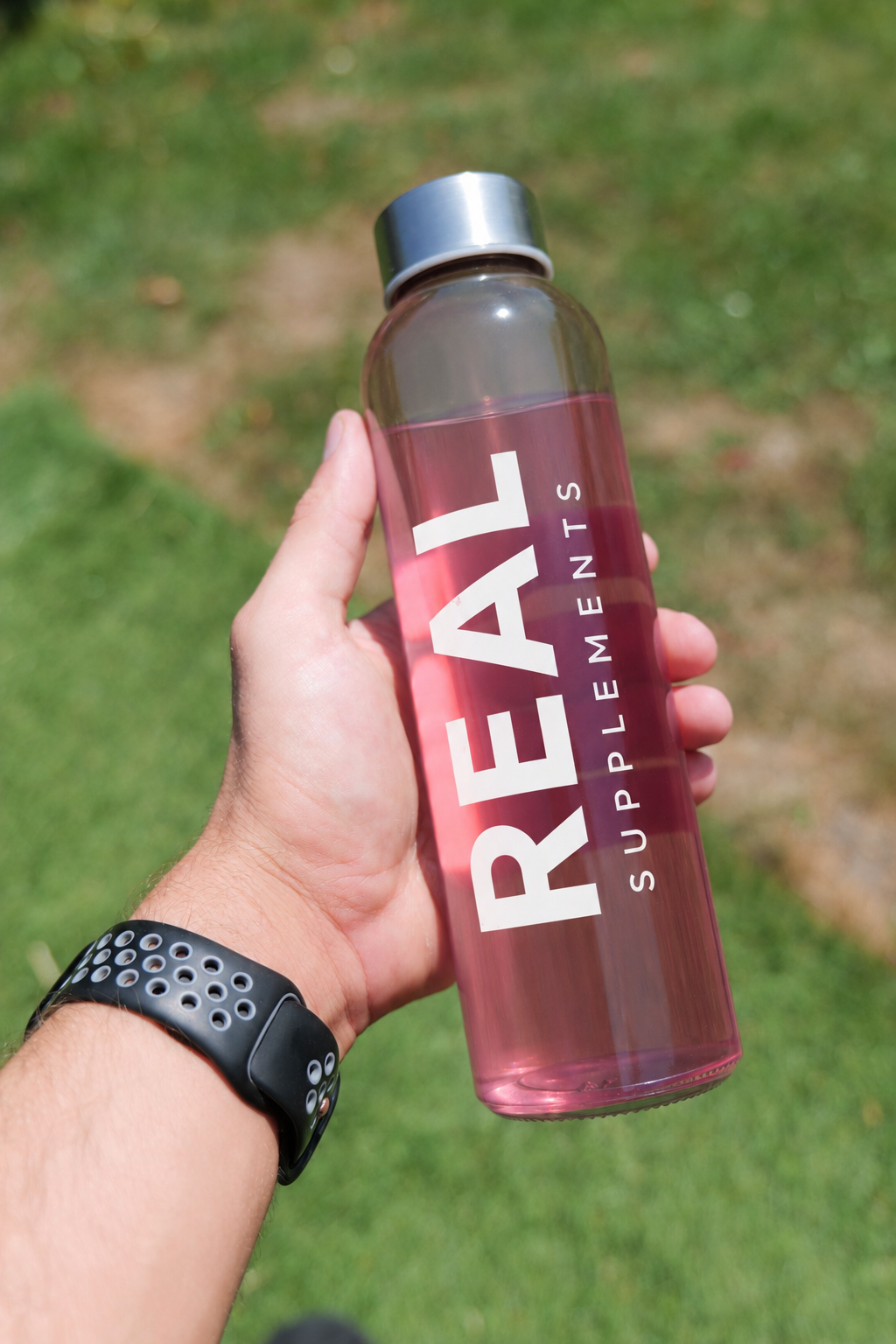 A hand holding a translucent pink water bottle with a silver cap, featuring the brand name 'REAL SUPPLEMENTS' printed in bold white letters, set against a blurred green grass background.